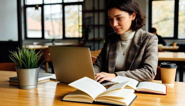 Young professional woman studying and working on laptop in cozy cafe with books and coffee