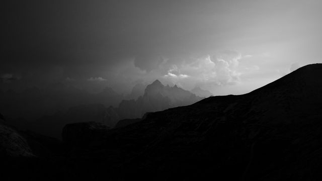 Panoramic view of a mountain range silhouetted against the dusk sky. Clouds billow in the distance, adding to the serene atmosphere. The black and white effect enhances the dramatic elements, making it ideal for use in artistic projects, meditation visuals, and atmospheric backgrounds.
