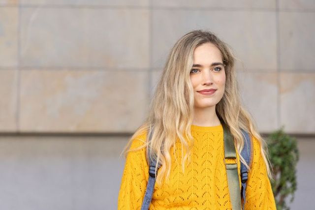 Young woman standing by tiled facade wearing yellow knit sweater and backpack