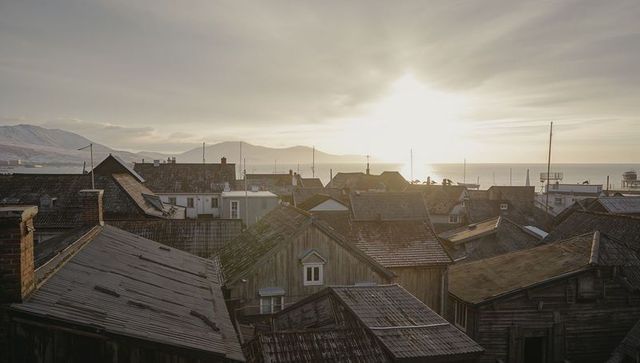 Weathered seaside rooftops catching golden sunrise with distant pier and mountains
