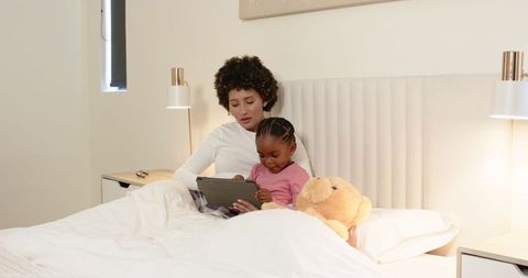 Mother and Daughter Relax in Bed Using Tablet with Teddy Bear