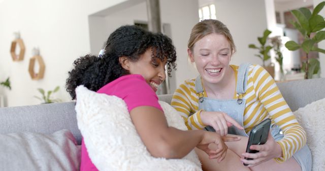 Women friends sitting on a couch, enjoying their time together. They are engaged in a conversation while looking at a smartphone, both smiling and laughing. This is perfect for depicting friendship, modern technology usage, and social media interaction. Ideal for use in ads for lifestyle products, social media apps, or articles related to friendship and social interactions.