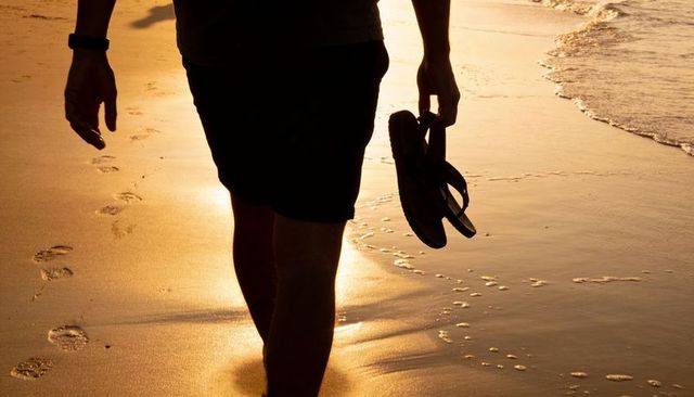 Silhouetted man walking barefoot along golden shoreline holding flip-flops at sunset