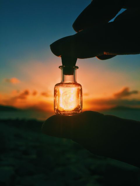 Hand holding small glass bottle while capturing beautiful sunset light by the beach creates a unique and artistic effect. Suitable for themes related to nature, serenity, creativity, and outdoor activities. Perfect for use in blogs, advertisements, art projects, posters, and travel content promoting relaxation and beauty of natural landscapes.