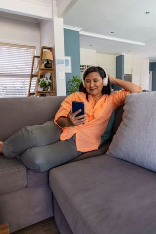 Mature asian woman reclining on sofa wearing headphones and using smartphone