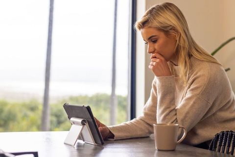 Remote professional woman in knit sweater working on tablet by window with coffee