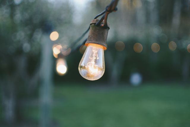 Outdoor vintage light bulb hanging on a string, glowing gently in twilight. Blurred bokeh background with greenery and another light. Ideal for use in content about outdoor decor, garden parties, cozy evening ambiance, or vintage aesthetics.