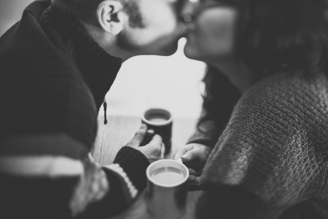 This image depicts a romantic couple sharing a kiss while holding mugs of coffee. They are dressed in cozy sweaters, creating a warm and intimate atmosphere. This can be used for promoting relationships, romantic holidays, coffee brands, lifestyle blogs, and Valentine's Day content.