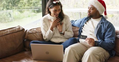 Diverse couple shopping online on laptop wearing santa hats sharing credit card on couch