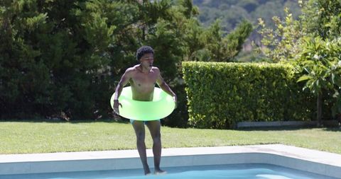 Man stepping into backyard pool holding neon green float on sunny lawn