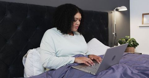 African American woman typing on laptop in cozy bedroom bed with tufted headboard and lamp