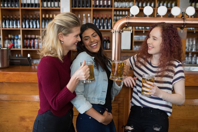 Three young women are standing at a bar counter, each holding a glass of beer and smiling. They appear to be enjoying each other's company in a casual and relaxed atmosphere. This image is perfect for use in advertisements for bars, pubs, or social events, as well as in articles or blogs about friendship, leisure activities, and social gatherings.