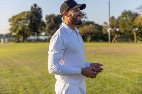 Smiling cricketer holding red ball on sunlit park field wearing white kit and black cap