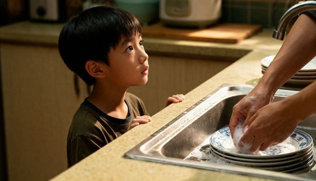 Young boy watching adult washing dishes at kitchen sink in warm home light, teaching