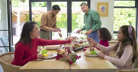 Family Gathering Around Sunlit Dining Table Sharing Holiday Meal and Passing Platters