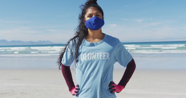 Portrait of biracial woman wearing volunteer t shirt and face mask looking at camera. eco conservation volunteers, beach clean-up during coronavirus covid 19 pandemic.