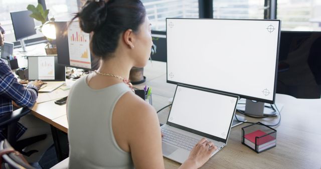 Businesswoman working on a laptop at a modern office desk with multiple monitors. Ideal for articles or marketing materials on productivity, workplace setup, technology in business, or corporate environments. Perfect for demonstrating a professional setting or modern office design.