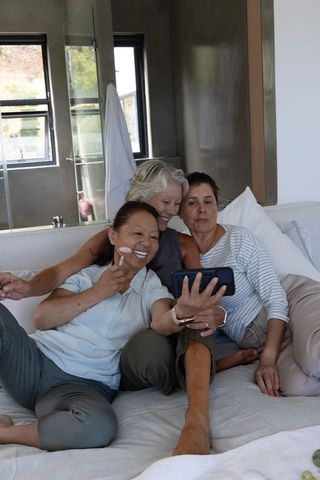 Mature diverse women taking selfie on bed, enjoying skincare and friendship in daylight