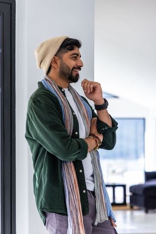Indian man leaning and smiling in minimalist living room wearing beanie and scarf