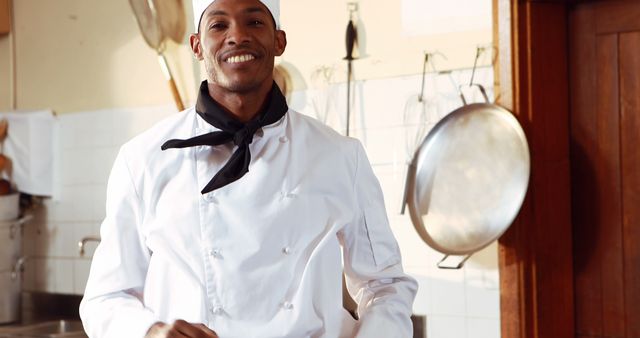 Male chef standing in professional kitchen, smiling. The chef is wearing a traditional white uniform, chef hat, and a black scarf. Use this image to promote culinary classes, restaurants, chef training programs, or cooking magazines. Showcasing professionalism and passion for cooking.