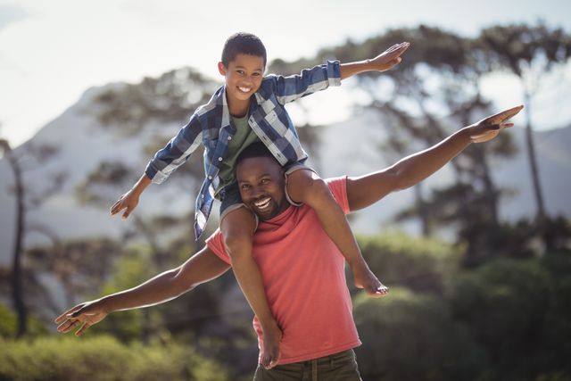 Father carrying his son on his shoulders, both extending their arms and smiling joyfully. Set against a backdrop of trees and mountainous terrain under a clear sky. Ideal for illustrating concepts of family bonding, parenting, happiness, and outdoor activities.