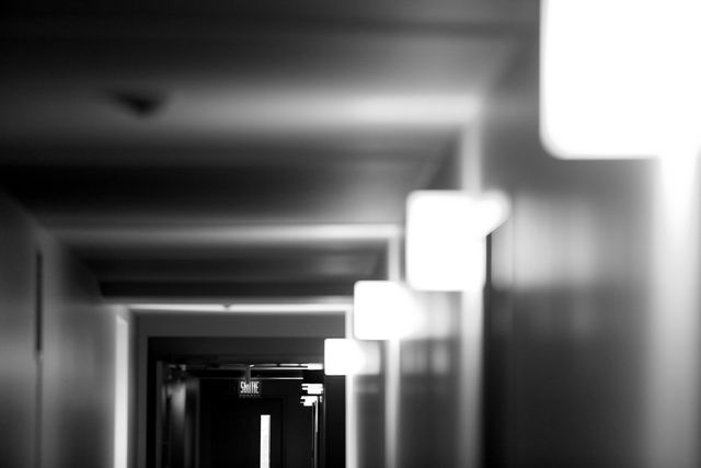 Black and white image of a dimly lit hallway with square light fixtures on the wall. The perspective draws the eye towards the exit at the end of the corridor, adding a sense of depth and mystery. This can be used in projects that require a mysterious or eerie atmosphere, perfect for backgrounds, presentations, or design elements in creative projects.