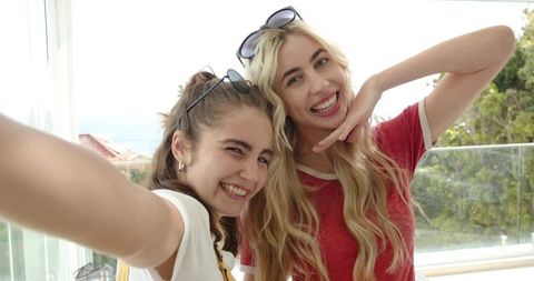 Two young women smiling and posing for selfie on sunny balcony overlooking ocean