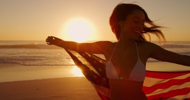 A joyful woman is holding the American flag while standing on a beach during sunset. She is wearing a white bikini and appears to be celebrating with a bright smile. The ocean waves and sunlight create a warm, vibrant atmosphere. This image is perfect for advertising beach resorts, summer vacations, patriotic events, or lifestyle blogs focusing on travel and adventure.