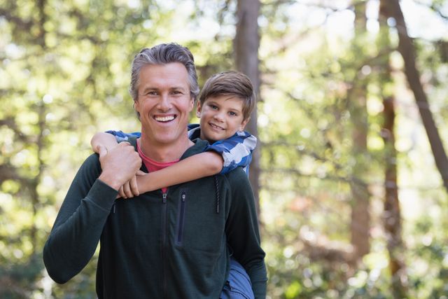 Father giving son a piggyback ride while hiking in a forest. Both are smiling and enjoying the outdoors. Ideal for use in family-oriented advertisements, parenting blogs, outdoor activity promotions, and nature-themed content.