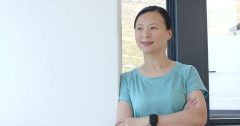 Asian woman leaning by window, wearing smartwatch, modern minimalist sunlit interior