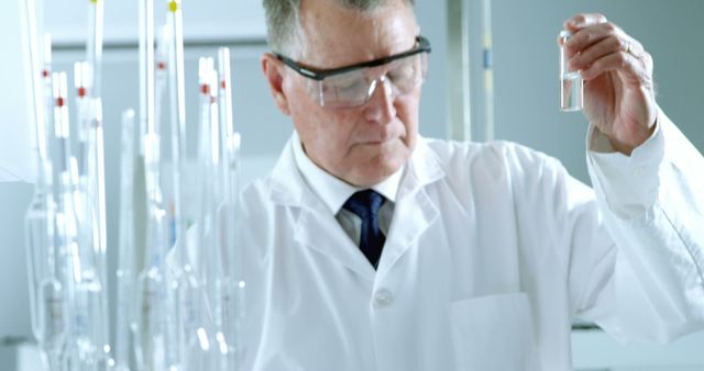 The image shows a senior scientist wearing safety glasses and a white lab coat while analyzing a sample in a laboratory. He is surrounded by laboratory equipment, emphasizing a professional research environment. This image is ideal for use in articles and presentations about scientific research, medical advancements, chemistry education, healthcare developments, and biotechnology innovations.