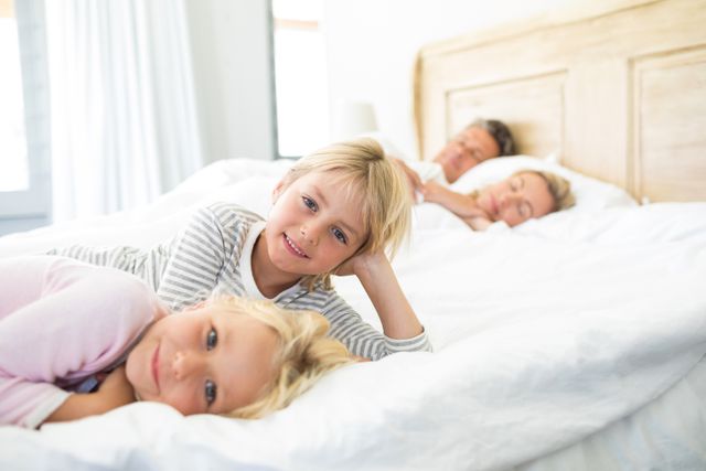 Two smiling children lying on a bed with their parents sleeping in the background. Perfect for family-oriented content, advertisements for home products, or articles about family bonding and morning routines.