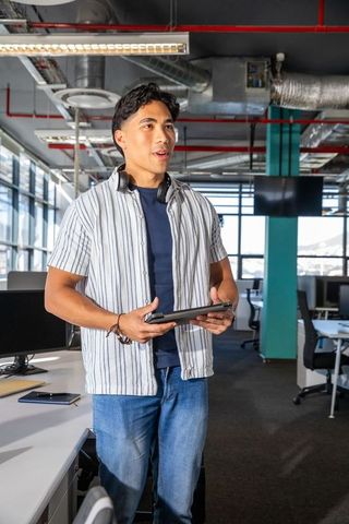 Asian man standing in modern open plan office holding tablet and wearing headphones