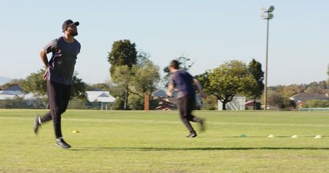 Coach leading diverse male team running agility drills across cones on green field fitness