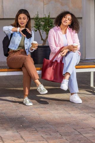 Two african american women checking smartphone and drinking coffee on urban bench