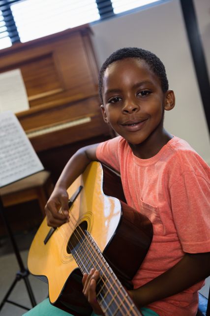 Young boy holding guitar and smiling in a music classroom. Ideal for educational materials, music school promotions, and articles on children's music education. Perfect for illustrating concepts of learning, talent development, and musical practice.