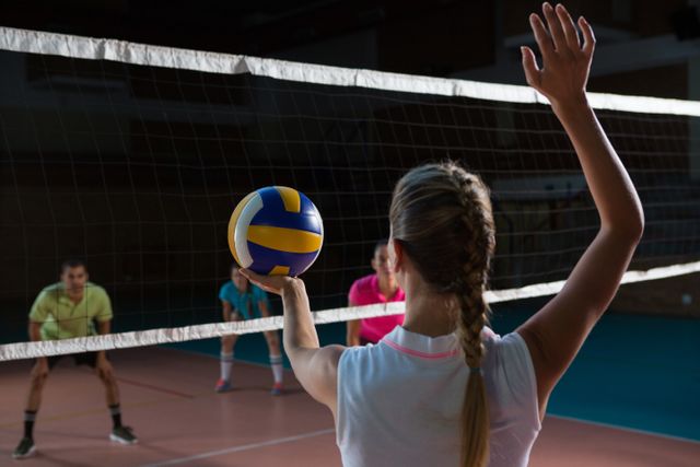 This image shows a female volleyball player preparing to serve the ball in an indoor court, with teammates ready on the other side of the net. Ideal for use in sports-related articles, fitness blogs, team-building promotions, and athletic training materials.