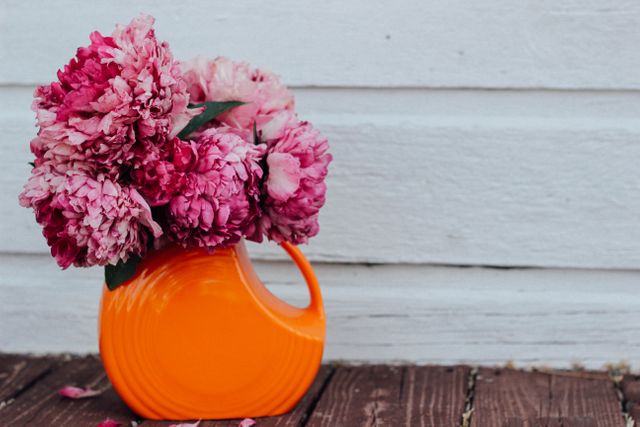 Pink flowers placed in an orange vase sitting on a rough wooden surface in front of a plain white wall. Perfect for use in home decor blogs, floral design websites, and social media posts celebrating nature and beauty.