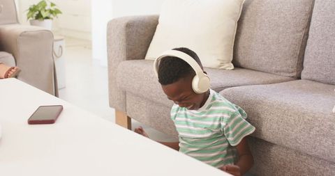 Smiling boy wearing white headphones sitting on living room floor beside gray sofa and smartphone
