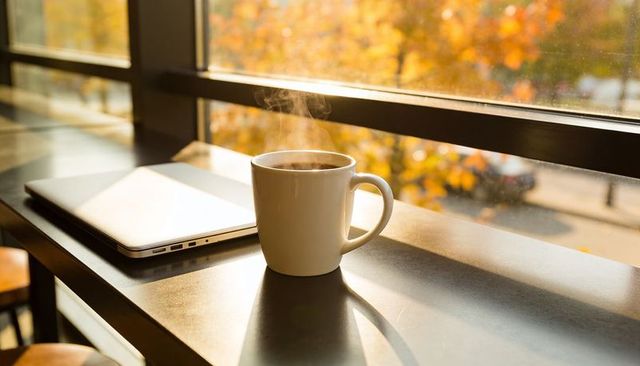 Steaming coffee mug by laptop on sunlit window counter in cozy autumn workspace