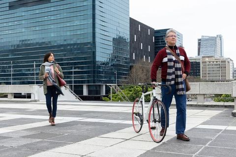 Senior man walking bicycle through modern glass plaza while woman carrying coffee cup