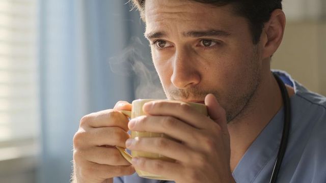 Doctor sipping steaming coffee while gazing out window in blue scrubs with stethoscope