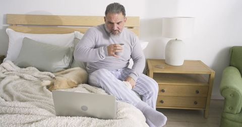 Senior man relaxing in bed with laptop and sleeping dog