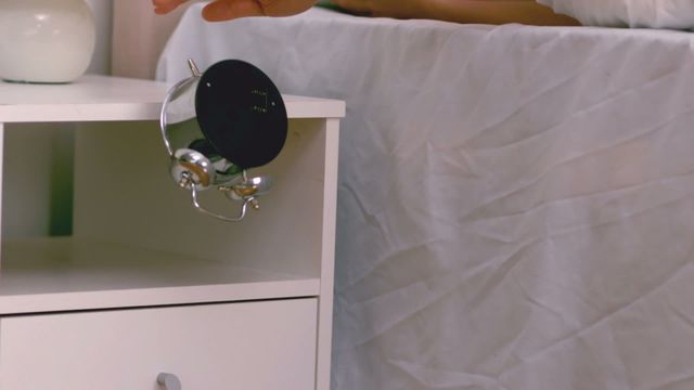 Hand pushing alarm clock off bedside table, illustrating difficulty waking up on a lazy morning; useful for themes about morning routines, alarm systems, waking up challenges, and sleep behaviors.