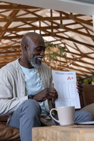 Mature african man smiling holding a+ report card in cozy wood-beamed living room