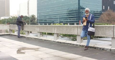 African american commuter leaning on overpass checking smartphone while eating sandwich