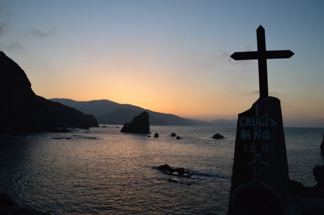 Cross with inscriptions stands on a cliff overlooking the ocean at sunset, with serene mountains in the distance. Useful for themes of spirituality, natural beauty, solitude, and tranquility.