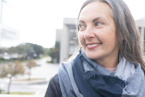 Mature woman smiling and gazing left on urban walkway wearing woven scarf and dark coat