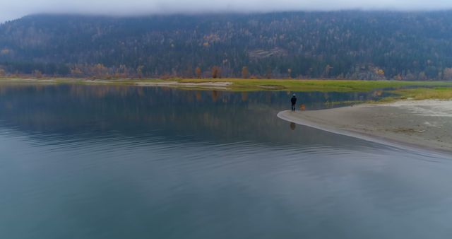 A solitary person is seen walking along a serene lake shore, with fog-shrouded mountains in the background. The reflection of the trees and mountains highlights the calm nature of the scene. This image can be used for themes of solitude, peace, and nature. It is ideal for websites, blogs, and marketing materials promoting travel, relaxation, and environmental conservation.