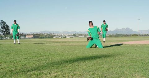 Baseball teammates kneeling and celebrating catch on sunlit field with mountain backdrop